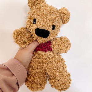 May include: A handmade teddy bear with a fuzzy, light brown body, black eyes, and a black nose. The bear has a red heart-shaped patch on its chest. The bear is being held up against a white background.