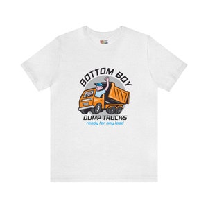 May include: White t-shirt featuring a cartoon graphic of an orange dump truck with a person waving from the driver's seat. The text "BOTTOM BOY" is above the truck, and "DUMP TRUCKS ready for any load" is below.