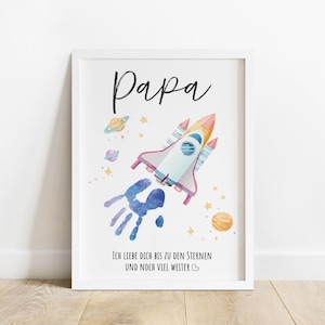 Könnte beinhalten: Ein gerahmter Druck mit einer Aquarellillustration einer Rakete, Planeten und Sternen. Das Wort "Papa" steht in Schreibschrift oben, darunter ein Handabdruck. Der Text "Ich liebe dich bis zu den Sternen und noch viel weiter" steht unten.