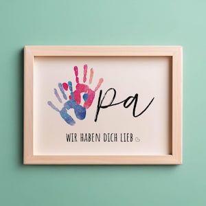 Peut inclure: Une impression encadrée avec deux empreintes de mains colorées et le mot "Pa" en écriture cursive noire. Le texte sous les empreintes de mains dit "Wir haben dich lieb" avec un petit symbole de cœur.