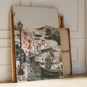 May include: A framed print of a coastal town with colorful buildings, boats, and a waterfront scene. The artwork features a gold frame and a vintage aesthetic, leaning against a white wall.