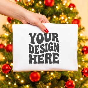May include: A white rectangular fabric item featuring the dark gray text YOUR DESIGN HERE in a bold, wavy, retro-inspired font. The text is centered on the front surface of the material, which is held up against a blurred holiday tree background.