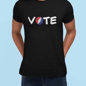 May include: Black t-shirt with the word "VOTE" in white, featuring a red and blue graphic. The graphic resembles a lightning bolt within a circle. The shirt is a crew neck style.