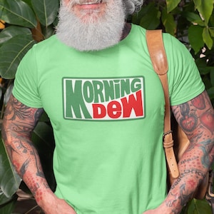 May include: A light green t-shirt with the text "MORNING DEW" in a green and red rectangular design. The person is wearing blue jeans and has tattoos on their arms.