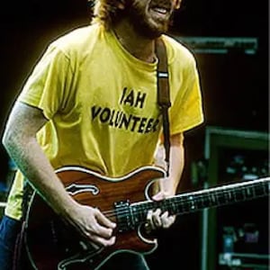 May include: A musician playing a brown electric guitar with a dark fretboard. The musician wears a yellow t-shirt with the text "IAH VOLUNTEER". The background is dark, suggesting a concert setting.