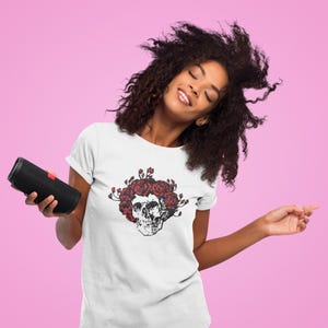 May include: White t-shirt featuring a black and red skull design with roses. The graphic tee has a crew neck and short sleeves. A black portable speaker is held in the left hand. The background is a solid pink color.