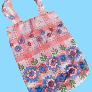 Puede incluir: Una bolsa de tela blanca con estampado floral rojo y azul. La bolsa tiene un patrón a cuadros y presenta flores azules con centros rojos y hojas verdes.