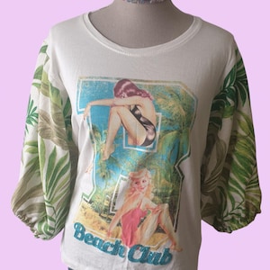May include: White t-shirt with a vintage beach scene graphic. The sleeves feature a green and white tropical leaf pattern. The graphic includes two women in swimsuits and the text "Beach Club".