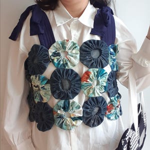Può includere: Un gilet unico, fatto a mano, con un design patchwork di cerchi in denim e tessuto floreale. Il gilet ha spalline e fiocchi blu navy sulle spalle. Il gilet è indossato sopra una camicia bianca abbottonata.