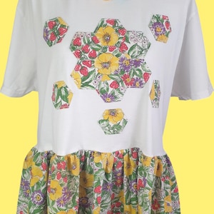 Puede incluir: Camiseta blanca con diseño floral y dobladillo con volantes. La camiseta presenta un patrón hexagonal de flores amarillas, fresas y flores moradas. Un collar de cuentas de colores está alrededor del cuello.
