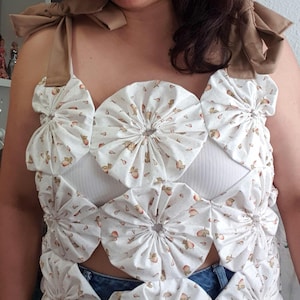 May include: A white crop top with a floral pattern of peaches and green leaves. The top features a unique design with gathered fabric circles and brown shoulder straps tied into bows. The top is paired with blue jeans.