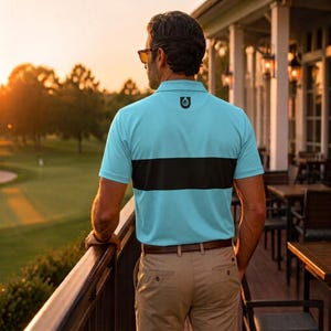 Man seen from behind at a clubhouse railing at sunset, wearing a Unanimous Golf light blue and black chest stripe polo with tan chinos, fairway in the background.