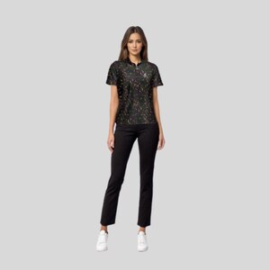 Full body shot of a female model wearing the black paint splatter golf polo, black pants, and white sneakers.