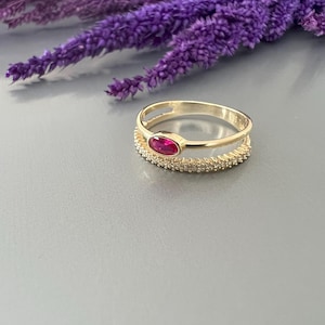 May include: A gold ring featuring a red oval gemstone set in the centre. The ring has a delicate, double band design with small clear stones.