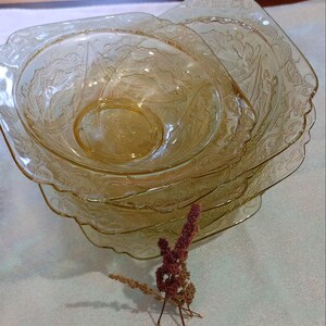 May include: A stack of four amber-colored glass bowls with a square shape and ornate, embossed floral designs. The bowls are nested, with a dried flower arrangement in the foreground. The bowls are likely vintage or antique.