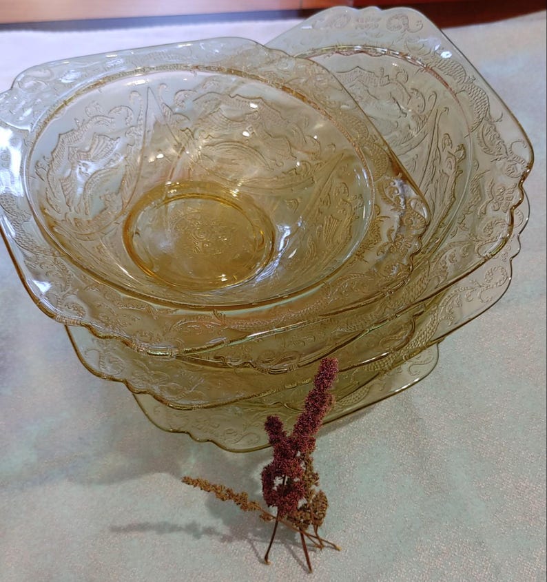 May include: A stack of four amber-colored glass bowls with a square shape and ornate, embossed floral designs. The bowls are nested, with a dried flower arrangement in the foreground. The bowls are likely vintage or antique.