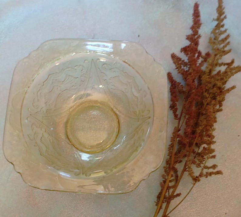 May include: A vintage, pale yellow glass bowl with a square shape and scalloped edges. The bowl features an etched floral design. A sprig of dried, reddish-brown flowers is next to the bowl.