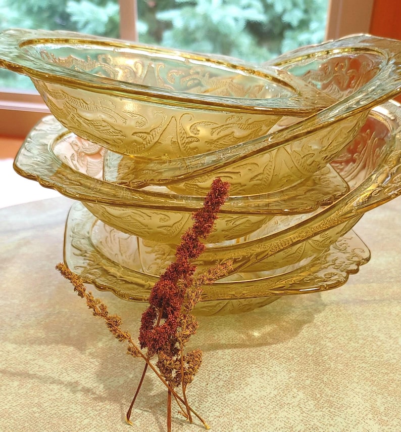 May include: A stack of amber-colored glass bowls with a floral design. The bowls have a scalloped edge and are stacked on top of each other. A few sprigs of dried flowers are in the foreground.