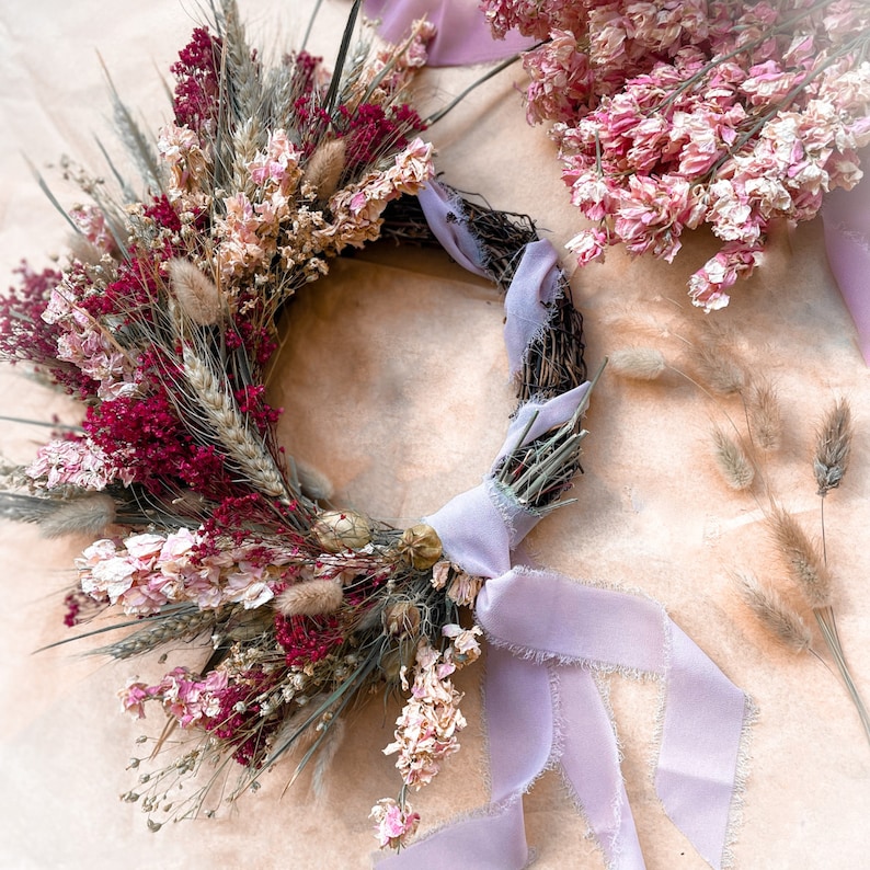 May include: A dried flower wreath with pink and burgundy flowers, wheat stalks, and a lavender ribbon bow.