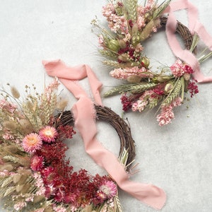 May include: Two dried flower wreaths with pink and red flowers, wheat, and grasses. Each wreath is decorated with a pink ribbon.