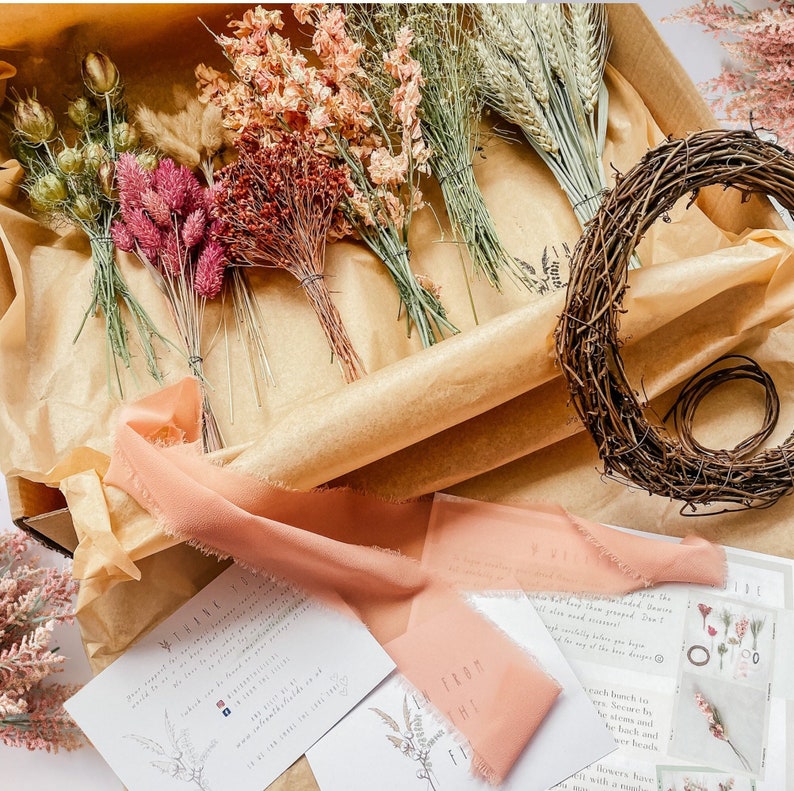 May include: A box filled with dried flowers and greenery, including pink, white, and green hues. A brown twig wreath is also included. The box is lined with brown paper and has a white card with the text "From the" on it.