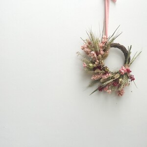 May include: A dried flower wreath with pink, white, and green flowers and greenery. The wreath is hanging from a pink ribbon.
