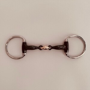 May include: A horse bit with a black center and silver rings. The bit has a gold-colored connector in the middle. The bit is designed for use in equestrian activities.