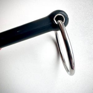 May include: A black equestrian bit with a silver metal ring. The bit is made of a dark material, and the ring is shiny and reflective. The design is simple and functional. The bit is likely used for horse riding.