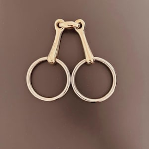 Könnte beinhalten: Ein Pferdgebiss mit einem goldfarbenen Mundstück und silberfarbenen Ringen. Das Gebiss ist mit zwei kreisförmigen Ringen entworfen, die durch einen goldfarbenen Steg verbunden sind. Die Ringe sind glatt und glänzend.