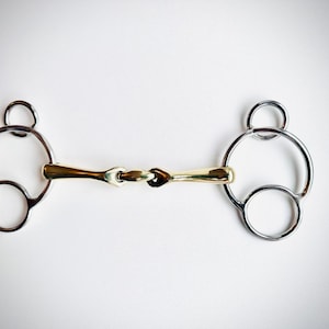 May include: A horse bit featuring a gold-coloured central section and silver-coloured rings. The bit has a jointed mouthpiece and circular rings on each side. The background is plain white.