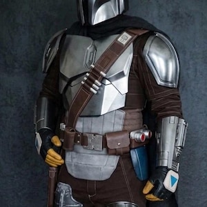 May include: A detailed costume of a Mandalorian warrior. The armor is primarily silver and gray, with brown and black accents. The helmet is a polished silver, and the outfit includes a belt with pouches and a holster. The overall aesthetic is futuristic and battle-ready.