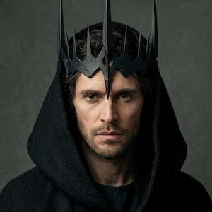 May include: A black crown featuring tall, sharp, pointed spikes arranged in a jagged, angular formation. The crown is worn over a dark, textured hooded garment that covers the head and shoulders, creating a stark, monochromatic appearance.