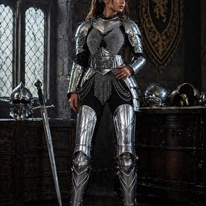 May include: A full suit of polished silver armor, including a breastplate, greaves, and gauntlets, is worn over a black outfit. A chainmail shirt and a sword are also visible. The setting is a medieval-style room with a large window.