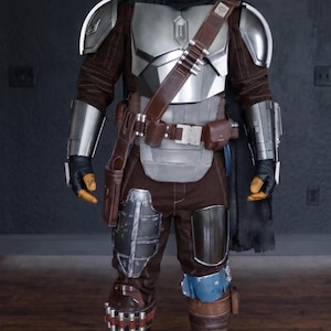 May include: A full-body shot of a Mandalorian costume. The armor is primarily silver and brown, with a helmet, chest plate, shoulder pads, and arm guards. The costume includes a brown leather belt, pouches, and a black cape. The boots are brown.