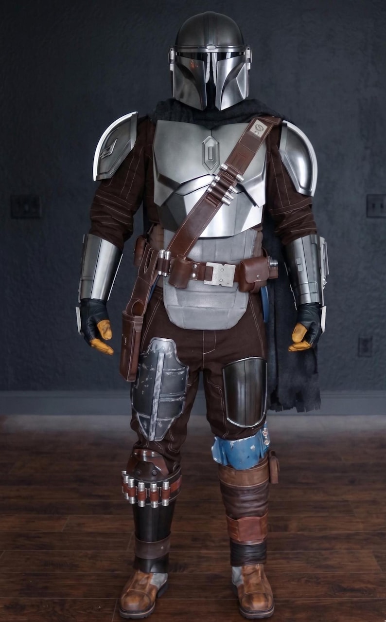 May include: A full-body shot of a Mandalorian costume. The armor is primarily silver and brown, with a helmet, chest plate, shoulder pads, and arm guards. The costume includes a brown leather belt, pouches, and a black cape. The boots are brown.