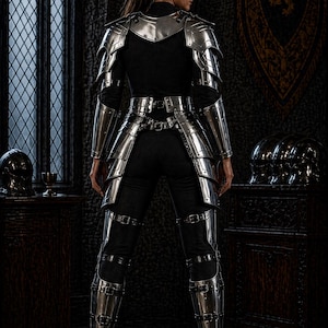 May include: A person wearing polished silver armor, including a breastplate, shoulder guards, arm guards, and leg armor. The armor is worn over a black top and pants. The background includes a dark room with a window and a decorative tapestry.