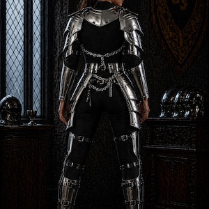 May include: A person wearing silver metal armor, including a chest plate, arm guards, and leg armor, over a black outfit. The armor is detailed with buckles and chains. The background features a dark interior with a window and decorative elements.