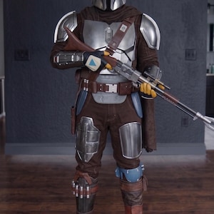May include: A full-body shot of a Mandalorian costume. The costume features a silver helmet, chest armor, and shoulder pads. The brown and silver outfit includes a blaster rifle, brown pants, and boots. The costume is detailed and appears to be a replica.