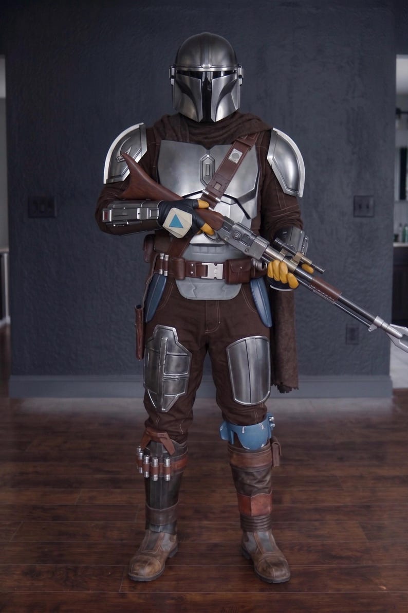 May include: A full-body shot of a Mandalorian costume. The costume features a silver helmet, chest armor, and shoulder pads. The brown and silver outfit includes a blaster rifle, brown pants, and boots. The costume is detailed and appears to be a replica.