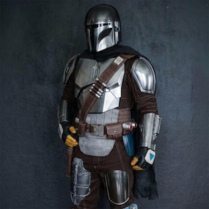 May include: A detailed costume of a Mandalorian warrior. The armor is primarily silver and brown, with a helmet, chest plate, and shoulder pads. The costume includes a brown leather belt, pouches, and a black cape. The gloves are yellow and black.
