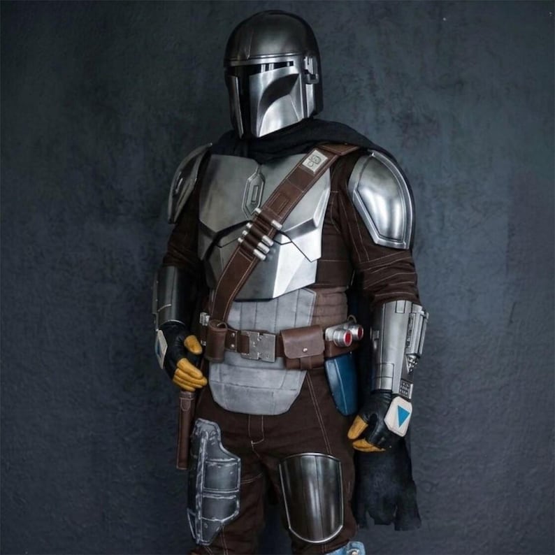 May include: A detailed costume of a Mandalorian warrior. The armor is primarily silver and brown, with a helmet, chest plate, and shoulder pads. The costume includes a brown leather belt, pouches, and a black cape. The gloves are yellow and black.