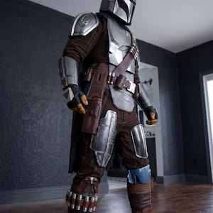 May include: A full-body shot of a Mandalorian costume. The costume features a silver helmet, chest plate, and arm guards over a brown and gray jumpsuit. Brown leather accents include a belt, holster, and boot details. The costume is designed with a futuristic aesthetic.