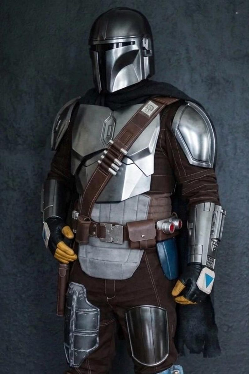 May include: A detailed costume of a Mandalorian warrior. The armor is primarily silver and gray, with brown and black accents. The helmet is a polished silver, and the outfit includes a belt with pouches and a holster. The overall aesthetic is futuristic and battle-ready.