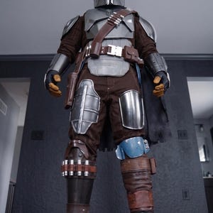 May include: A full-body shot of a Mandalorian costume. The costume features a silver helmet, chest armor, and shoulder pads. Brown and gray clothing is visible beneath the armor. The costume includes a brown belt, pouches, and boots. A black cape hangs from the back.