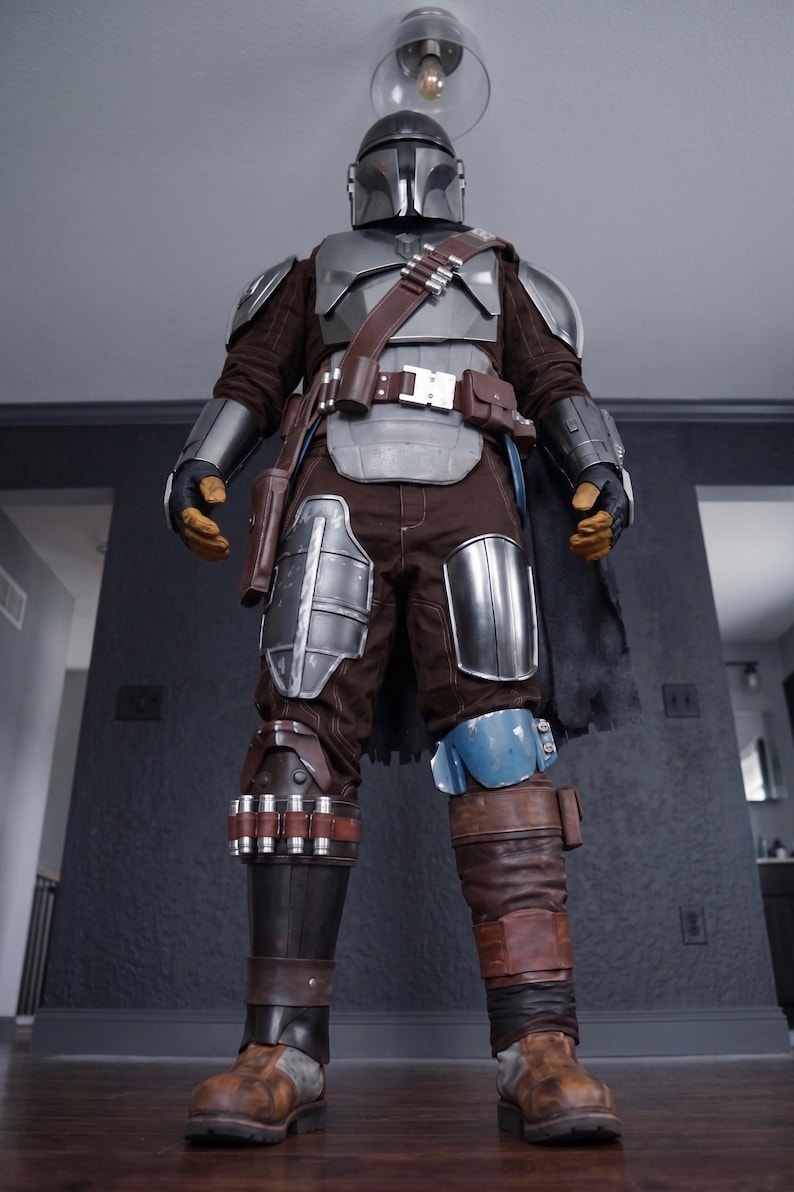 May include: A full-body shot of a Mandalorian costume. The costume features a silver helmet, chest armor, and shoulder pads. Brown and gray clothing is visible beneath the armor. The costume includes a brown belt, pouches, and boots. A black cape hangs from the back.