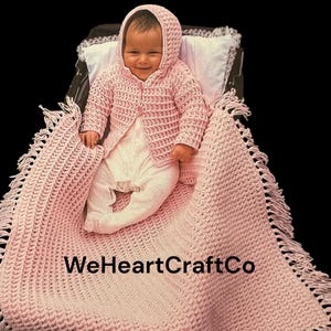 Può includere: Un bambino con una giacca con cappuccio rosa all'uncinetto e una coperta abbinata. La coperta ha un bordo con frange e il testo "WeHeartCraftCo" è visibile. Il bambino è sdraiato su un cuscino bianco.