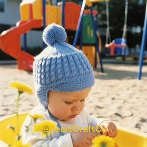 May include: A light blue knitted baby hat with ear flaps and a pom-pom. The hat is worn by a baby. The text "We Heart Craft Co" is visible at the bottom of the image.