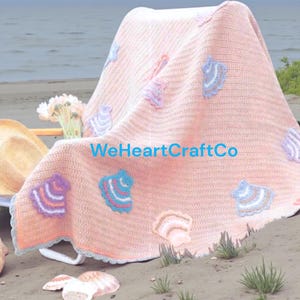 May include: A peach-colored crochet blanket with seashell appliqués in various colors, draped over a chair on a sandy beach. The blanket has a scalloped edge and the text "WeHeartCraftCo" is visible.