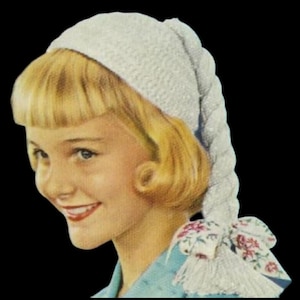 May include: A vintage-style white knitted cap with a long, braided tail and a floral bow. The cap is worn by a person with blonde hair and a blue top. The bow features a pink and red floral pattern.