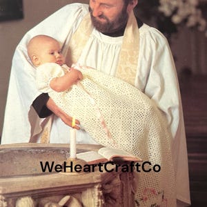 May include: A baby in a white crocheted dress and blanket is held by a person wearing a white robe. A lit candle and an open book are on a stone baptismal font. The text "WeHeartCraftCo" is visible.